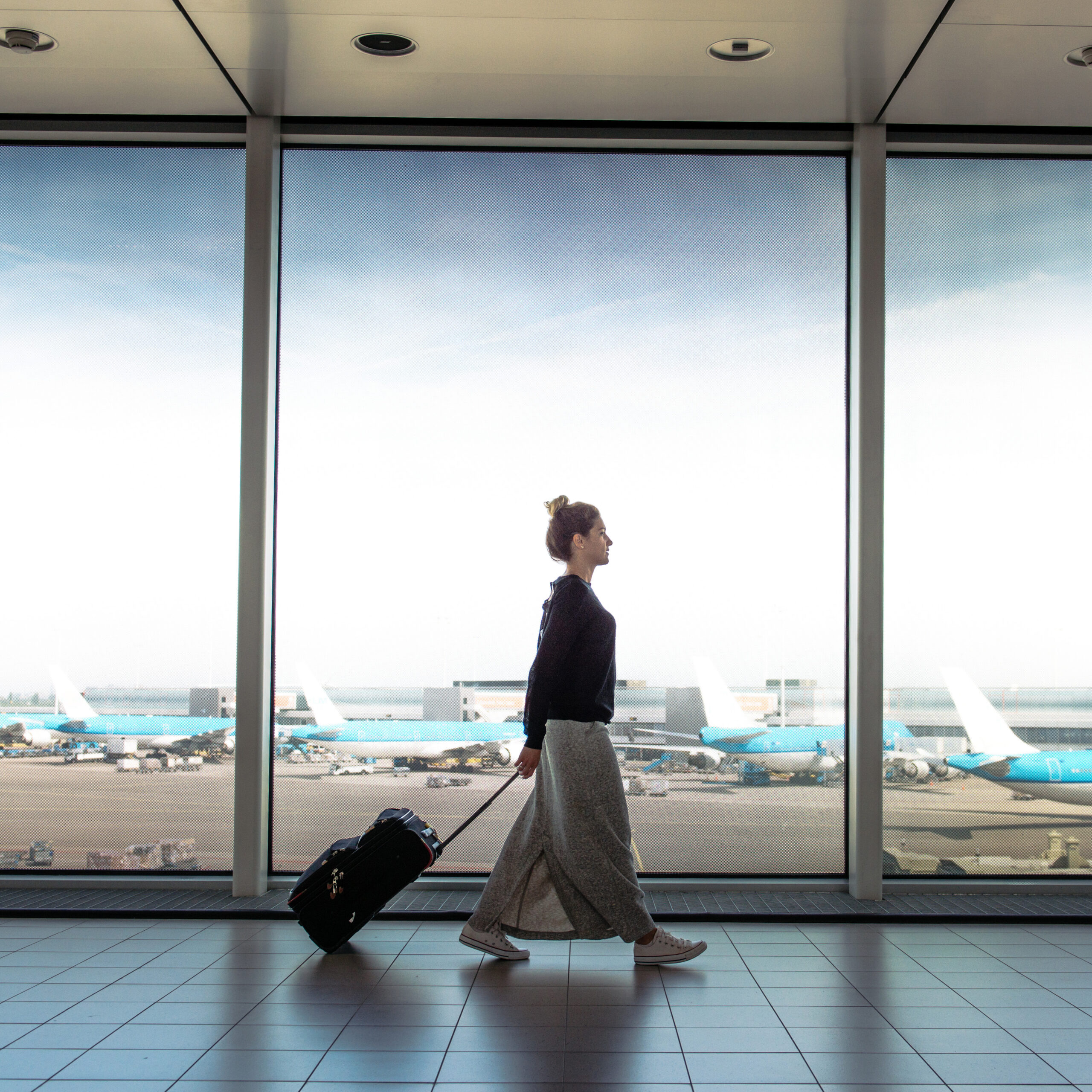 Frau läuft mit einem kleinen Koffer an einem Flughafen eine Fensterfront entlang