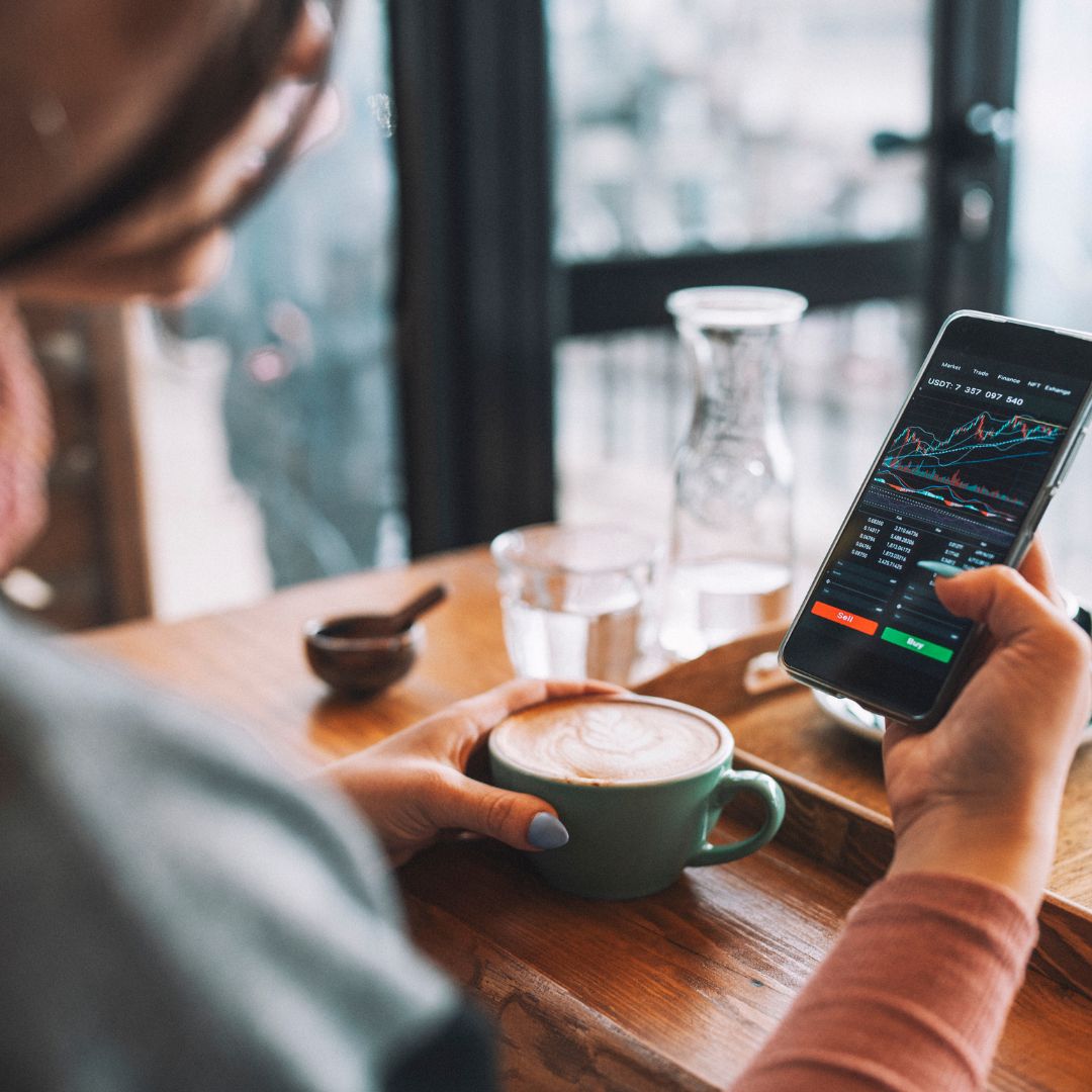 Eine Frau sitzt in einem Cafe und hat in der einen Hand eine Kaffee-Tasse und in der anderen Hand ein Smartphone. Auf dem Smartphone sieht man Kursentwicklungen.