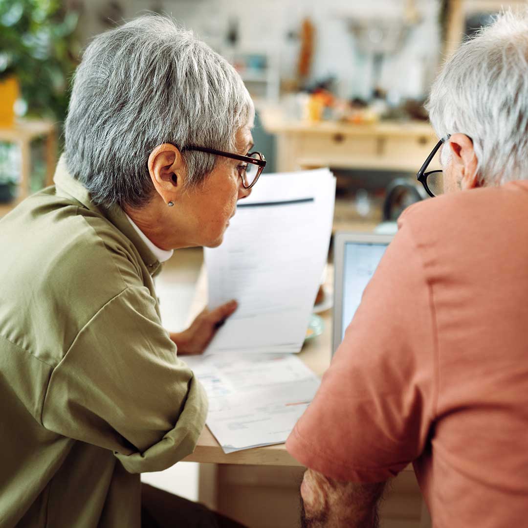 Eine ältere Frau und ein älterer Mann sitzen gemeinsam an einem Tisch und sehen sich Dokumente an.
