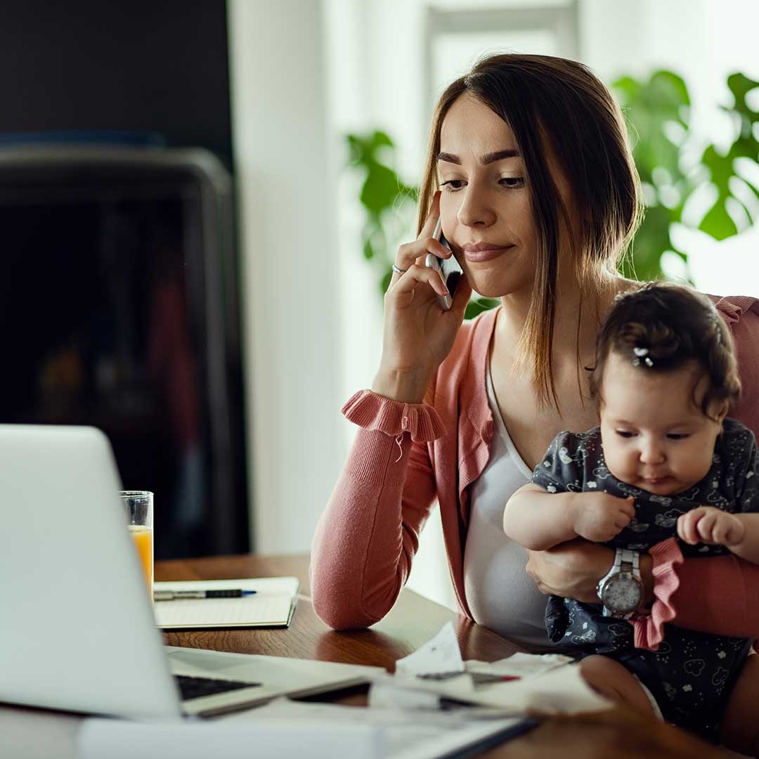 Berufstätige Frau sitzt mit Kleinkind im Arm im Home Office