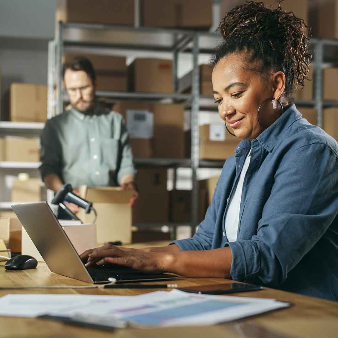 Eine Frau mit lockigen hochgesteckten Haaren sitzt an einem Tisch und tippt auf einem Laptop. Im Hintergrund steht ein Mann der vor einem Lager mit Pappkartons ein Paket verpackt.