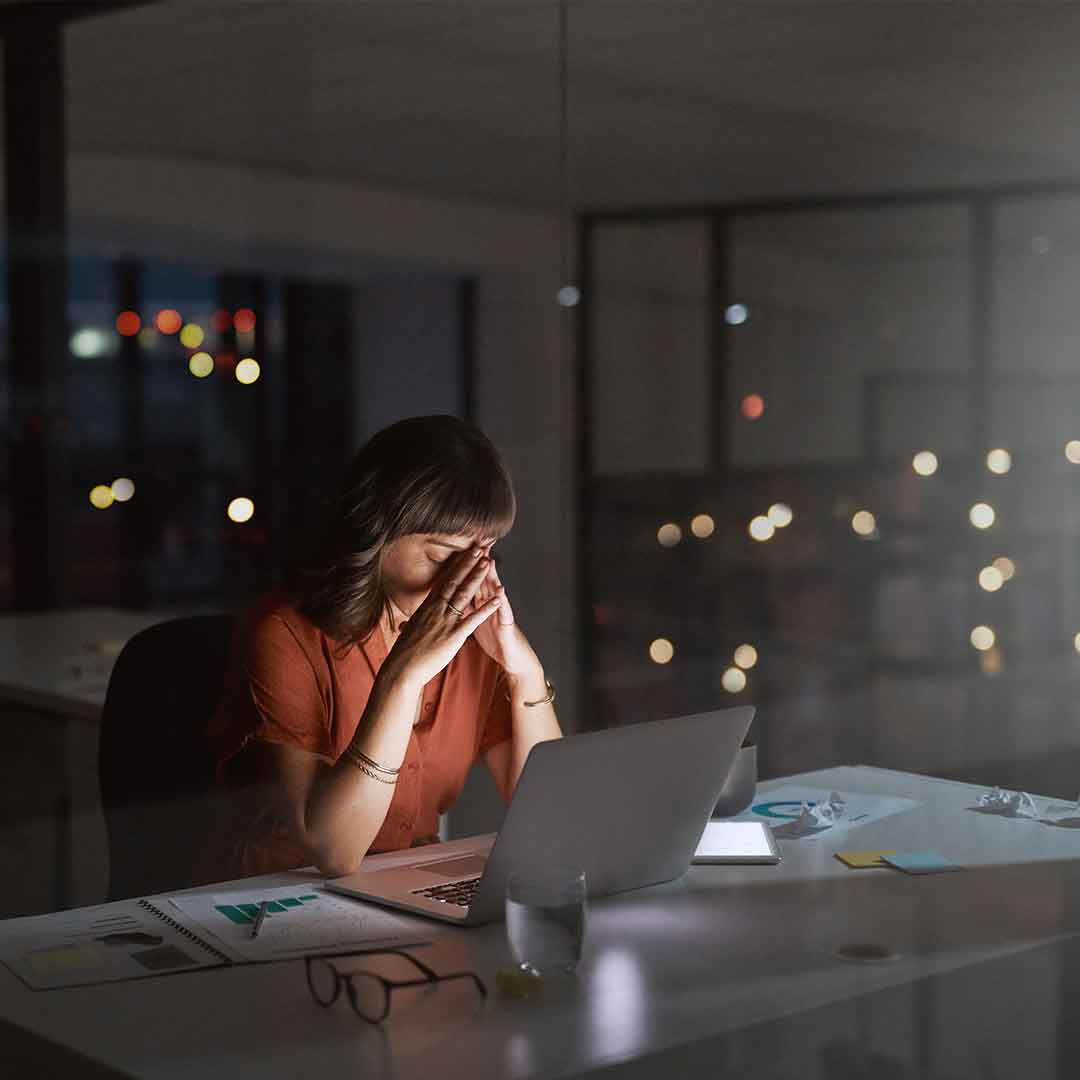 Eine übermüdete und gestresste Frau sitzt spät abends vor ihrem Laptop und schlägt die Hände vor dem Gesicht zusammen